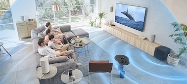 Family on a sofa watching TV with the HT-A9 Home Theatre System on a wooden cabinet with sound  waves emanating from the subwoofer.