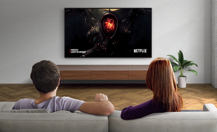 Image of a couple in a living room watching Netflix on a wall-mounted BRAVIA TV. When the Adaptive Calibrated Mode button below the image is toggled on and off, you can see the screen brightness change to match the environment.